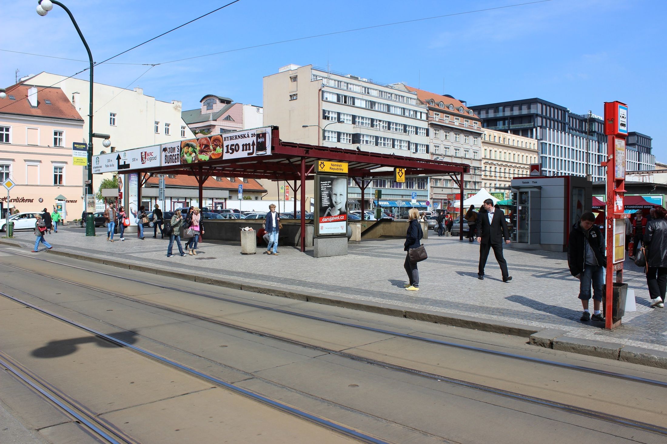 Pohled z protějšího chodníku na prostor - výstup z metra před Masarykovým nádražím. Vpravo je vidět označník tramvajové zastávky směr Bílá labuť nebo Náměstí Republiky.